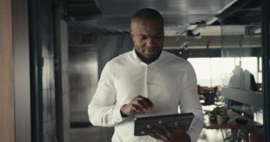 Kendine güveni tam bir siyahi modern ofiste yürüyor ve bir tablet üzerinde çalışıyor. Ofiste takım elbiseli bir adam yürüyor..