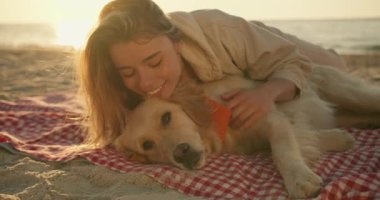Yakın çekim: Sarışın bir kız ve parlak renkli şirin köpeği sabahları güneşli bir plajda yan yana yatıyorlar. Evcil hayvanınla iyi vakit geçir..