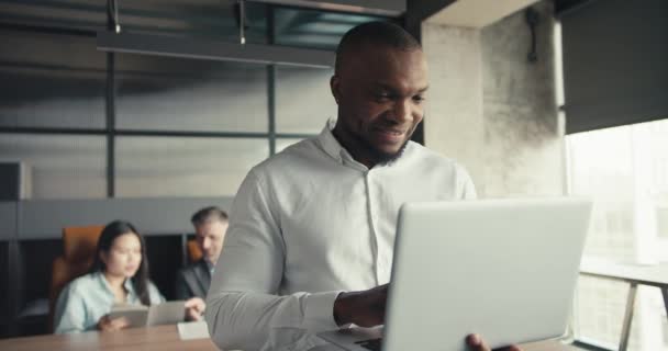 穿着白衬衫的自信的黑人在同事的背景下 在办公室的笔记本电脑上工作 办公室里的团队精神 通向成功的道路 — 图库视频影像