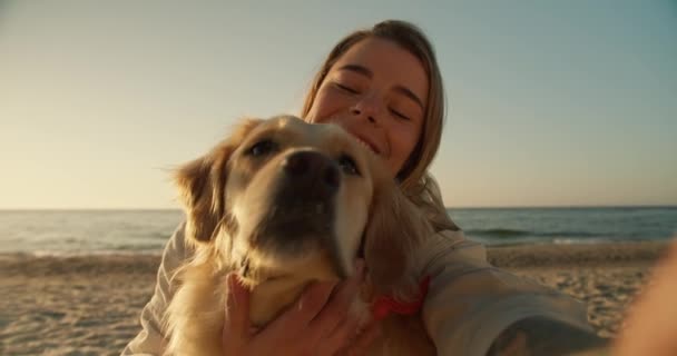 携帯電話の側からの眺めブロンドの女の子と彼女の軽い犬は 午前中に日当たりの良いビーチの背景に自撮りを取り クローズアップショット — ストック動画
