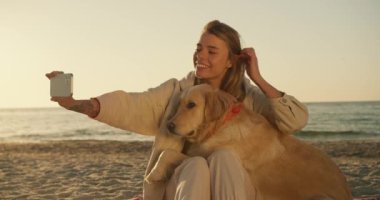 Bir kız, sabahları güneşli bir plajın arka planına karşı büyük şirin renkli köpeğiyle selfie çeker..