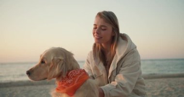 Sabahleyin deniz kıyısında köpeğini okşayan genç sarışın bir kızın portresi. Gün doğumunda sahilde bir evcil hayvanla sabah yürüyüşü.