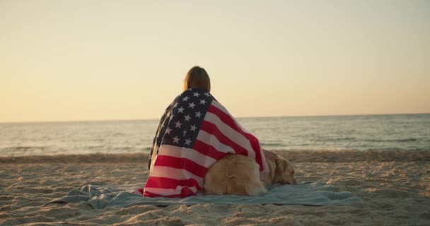 金发姑娘坐在阳光明媚的海滩上 她的狗裹着美国国旗 坐在阳光明媚的海滩上 — 图库视频影像