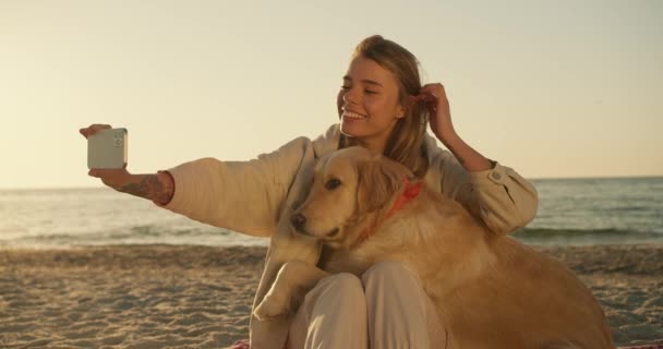 女の子は日当たりの良いビーチを背景に朝の光の色の彼女の大きなかわいい犬と一緒に自撮りを取ります — ストック動画