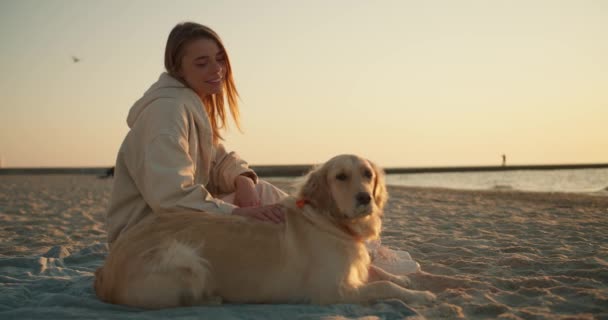 ブロンドの女の子は日当たりの良いビーチに座っています彼女の犬と一緒に明るい色で朝 — ストック動画