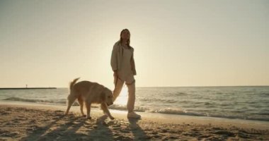 Sarışın bir kız, sabah güneşli bir sahilde açık renkli bir köpekle yürüyor..