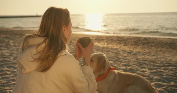 ブロンドの女の子は午前中に日当たりの良いビーチで魔法瓶のマグカップから熱いお茶を飲む 女の子の近くにある彼女の大きなかわいい犬の光の色 — ストック動画