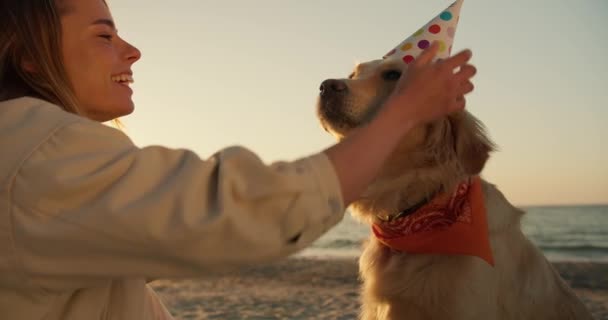 近身射击 一个金发女孩在阳光灿烂的海滩上祝她的狗生日快乐 并给它戴上节日的帽子 — 图库视频影像