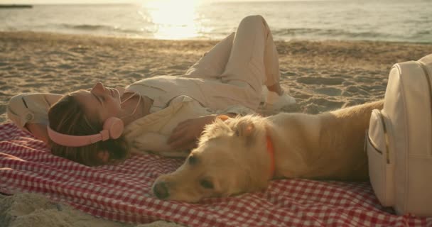 一位穿着浅色衣服的金发女孩一边用耳机听音乐 一边和她的狗躺在沙滩上晒太阳 — 图库视频影像