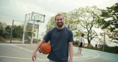 Gri tişörtlü, elinde basketbol topuyla basketbol sahasının dışında basketbol oynayan bir takımın arka planına poz veren kızıl saçlı bir adamın yakın plan fotoğrafı..