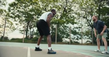 Beyaz tişörtlü siyah bir adam ve gri tişörtlü kızıl saçlı bir adam basketbol sahasında birbirlerine top atıyorlar. Kızıl saçlı adam topu basketbol potasına atmaya çalışıyor..