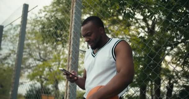 一个穿着白色T恤的黑皮肤小伙子笑了 手里拿着一个篮球 在智能手机上键入了一条信息 — 图库视频影像