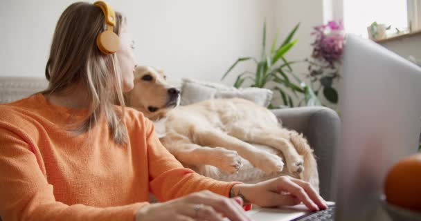 一个漂亮的金发女孩在笔记本电脑前工作 而她的狗却分散了她的注意力 但她们很快乐 在家里和你的宠物一起工作 — 图库视频影像