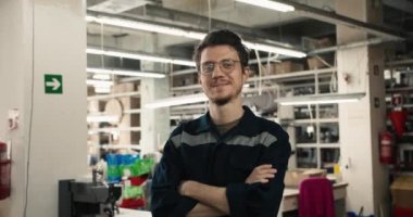 Gözlüklü ve mavi üniformalı bir adam gülümseyip tekstil fabrikasının önündeki kameraya bakıyor. Fabrika işçisi portresi.