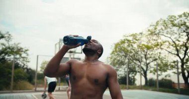 Siyah derili ve çıplak gövdeli bir adam basketbol sahasında su içer ve sonra serinlemek için kafasını yalar ve arkadaşlarının önünde bağırır..