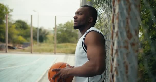 白いTシャツを着た若い黒人がバスケットボールコートのフェンスの近くに立っていて バスケットボールを手にしている アクティブな屋外レジャー スポーツ活動 — ストック動画