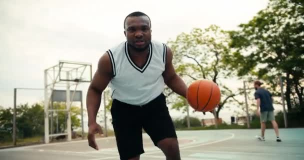 白いTシャツを着た黒人青年は 夏に外のバスケットボールコートでバスケットボールを巧みに扱う姿を見せてくれる — ストック動画