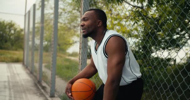 身穿白色T恤的疲惫的黑人手牵着篮球 站在室外运动场上 — 图库视频影像