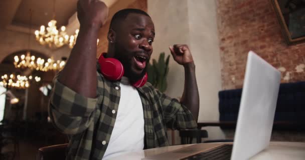 一个留着红色耳机胡子的黑人在咖啡店的笔记本电脑上工作 为成功感到高兴 并举起双手作为胜利的标志 在你最喜欢的工作中的成功情绪 — 图库视频影像