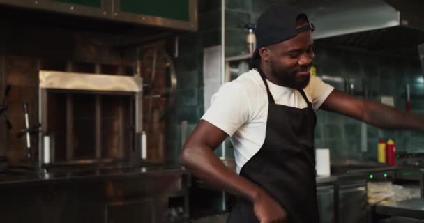 一个黑人在烤肉市场的背景下跳舞 穿着黑色围裙和白色T恤的人在工作中玩乐 — 图库视频影像