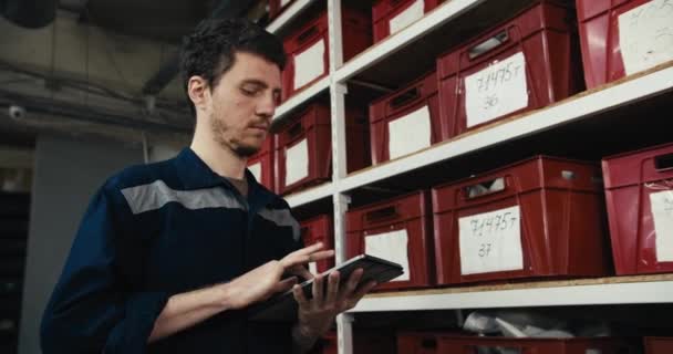 青い制服の赤い髪の男が靴工場で在庫を取ります タブレットの棚に赤い箱の中の靴の位置を書き留める男 — ストック動画