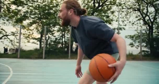 グレーのTシャツを着た赤い髪の男がオレンジのバスケットボールをドリブルします 相手はボールを奪おうとするが男たちは彼を守ろうとする — ストック動画