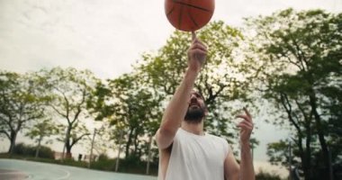 Beyaz tişörtlü orta yaşlı bir adam yazın bir spor sahasında işaret parmağında turuncu bir basketbol topunu ustalıkla çeviriyor..