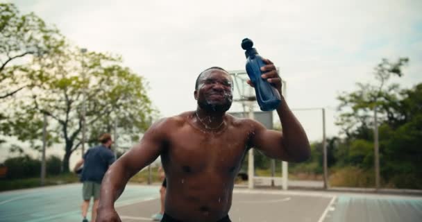 一个皮肤黑的人打篮球后 用一个特殊的瓶子把水倒入他的头上 冷却下来 然后在篮球场上大声喊着他的战斗口号 — 图库视频影像