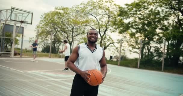 一个年轻的黑人男子的画像 他站在那里看着摄像机 在朋友们面前微笑 朋友们正在外面打篮球 — 图库视频影像