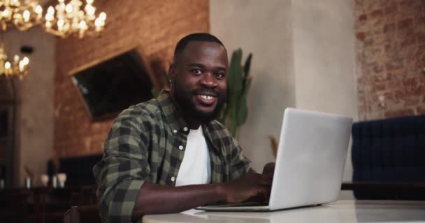 一个身穿格子绿色T恤的黑人坐在咖啡店的桌子旁 拿着笔记本电脑 看着相机笑了 — 图库视频影像