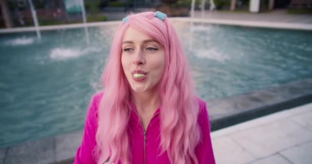 一个穿着粉色衣服的女孩正坐在喷泉边 一边玩乐 一边给泡泡糖气球充气 快乐而聪明的小女孩 — 图库视频影像
