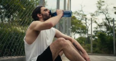 Beyaz tişörtlü orta yaşlı bir adam çitin yanındaki basketbol sahasında oturur, özel bir spor şişesinden su içer ve dinlenir..