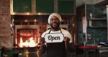 Özel bir başlık takmış bir siyah adam, bağış pazarındaki bir barbekü arka planında açık tabela ile poz verir..