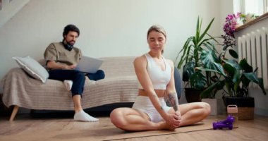 Güneşli bir odada bir çift: Kanepede oturan bir adam dizüstü bilgisayarda çalışırken sarışın bir kız özel bir halıda yoga yapıyor. Aynı odada çift etkileşimi..