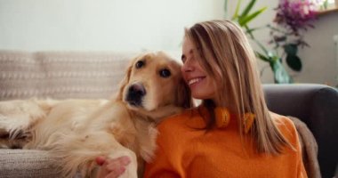Turuncu tişörtlü mutlu bir genç sarışın kız yere oturur ve kanepede oturan köpeğini okşar. Evcil hayvanınla mutlu vakit geçir..