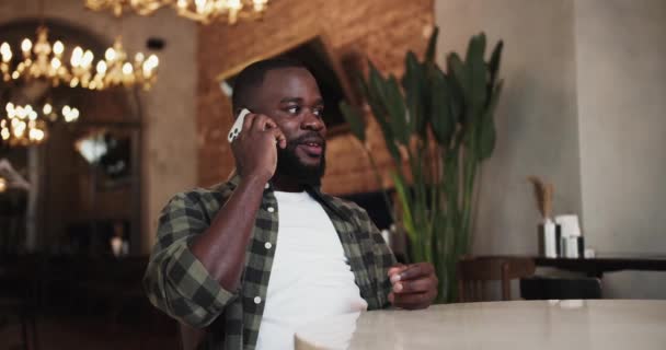 ポートレート チェック柄の緑色のTシャツを着た黒人がカフェのテーブルに座り 電話で話しています カフェでの電話での幸せな会話 — ストック動画