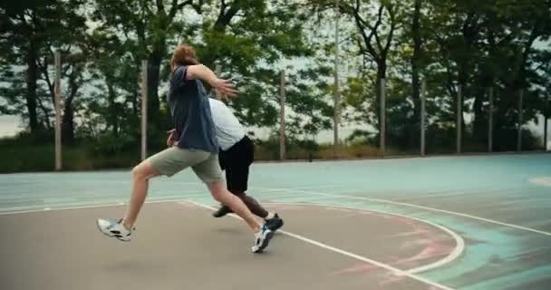 白いTシャツを着たバックマンとグレーのTシャツを着た赤髪の男がバスケットボールをしている 黒男は相手を追い越し バスケットボールの試合で素晴らしいゴールを決めました — ストック動画