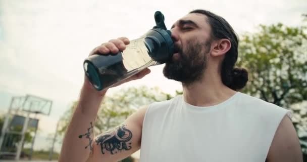腕にタトゥーのある中年の男と白いTシャツの男がバスケットボールコートで水を飲む — ストック動画