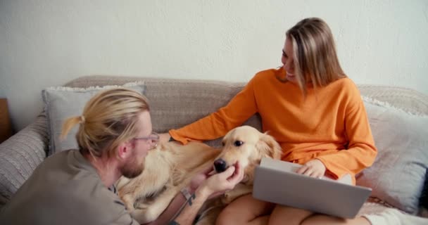 幸せなカップルで日当たりの良い部屋 ブロンドの男で眼鏡ペット犬 ブロンドの女の子はノートパソコンで動作し 犬に時間を捧げます — ストック動画