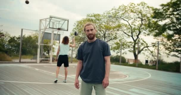 夏にバスケットボールコートでプレーするチームの前で 灰色のTシャツの赤い髪の男がポーズをとって胸に腕を折ります — ストック動画