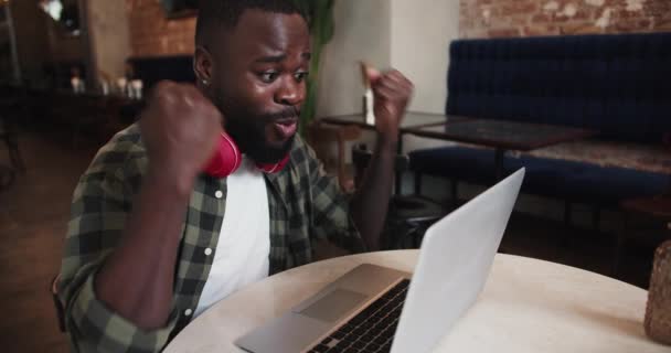 一个留着红色耳机和格子呢衬衫的留着胡子的男人在咖啡店的笔记本电脑上工作 为自己的成功感到高兴 并握紧拳头 — 图库视频影像
