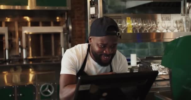 一个黑人在烤肉的背景下在烤肉市场上跳舞 一个穿着黑色围裙和白色T恤的男人对他最喜欢的工作感到高兴 — 图库视频影像