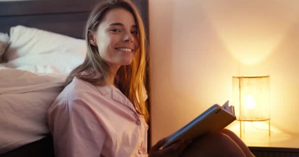 ブロンドの女の子はベッドの近くの床の上に座って本を読んでポーズし カメラを見ています 黄色のランプの近くの朝の読書本 — ストック動画