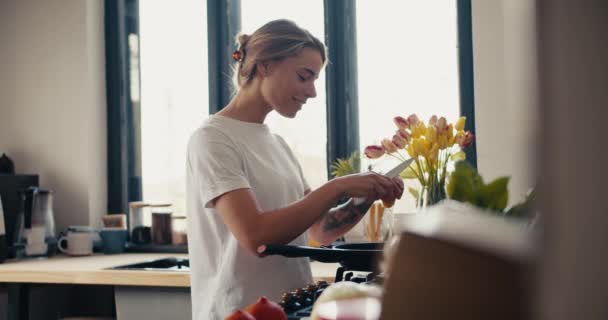 白いTシャツのブロンドの女の子は台所でスクランブルエッグを作ります 近くには花瓶の春の花 — ストック動画