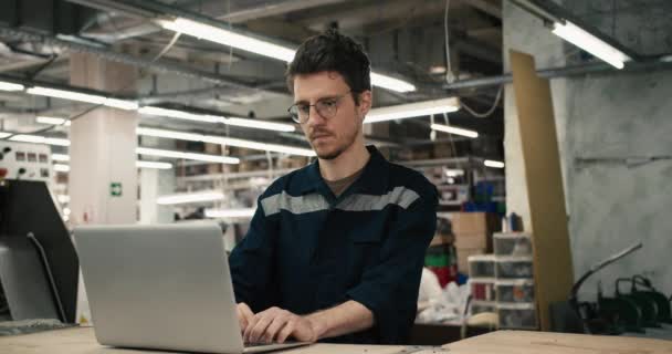 一名戴眼镜 身穿蓝色制服的男子在一家纺织厂的背景下 在一台白色笔记本电脑上工作 录像质量很高 — 图库视频影像