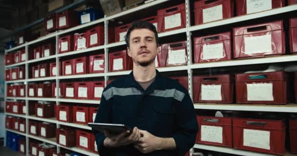 店内の倉庫で靴を履いた赤い箱を背景に タブレットを手にした青い制服の赤い髪の男がポーズをとって笑顔 高品質で撮影されたビデオ — ストック動画