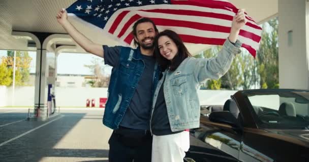 彼らの頭の上に米国の旗を上げ 彼らはガソリンスタンドで灰色のコンバーチブルの近くに立つ — ストック動画