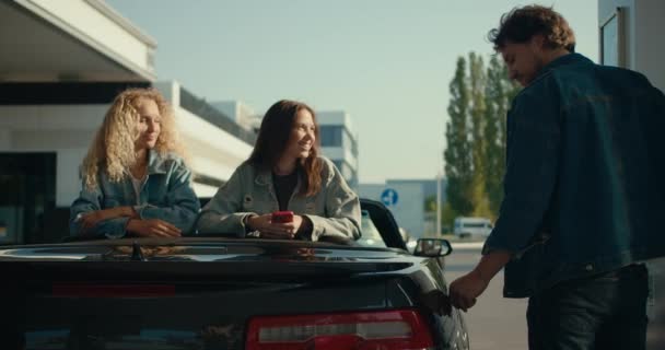 幸せな友人と灰色のコンバーチブルのグループ 2人の女の子は男が彼の車を埋めるのを見る 幸せな車に乗る ガソリンスタンドの停止 — ストック動画