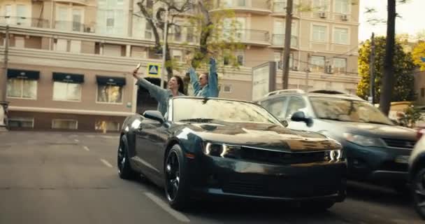 2人の幸せな女の子が楽しさを持って灰色のコンバーチブルに乗っていると自撮りを取ります 市内の幸せな若者たち — ストック動画