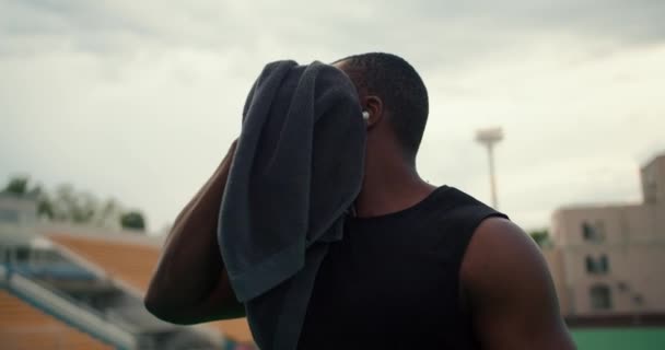 一名身穿黑色T恤 皮肤漆黑的运动员用毛巾擦拭自己的脸 并在城市海滩的灰蒙蒙的天空中喝着来自特殊瓶子的水 — 图库视频影像